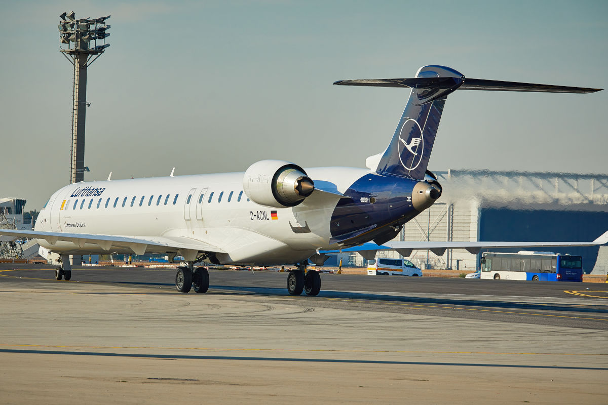 Auf dem Bild ist ein Flugzeug von Lufthansa Cityline auf dem Rollfeld eines Flughafens zu sehen, umgeben von Flughafenfahrzeugen und moderner Infrastruktur. Die Aufnahme zeigt das moderne Passagierflugzeug mit dem Lufthansa-Kranich-Logo am Leitwerk, bereit für den nächsten Flug.