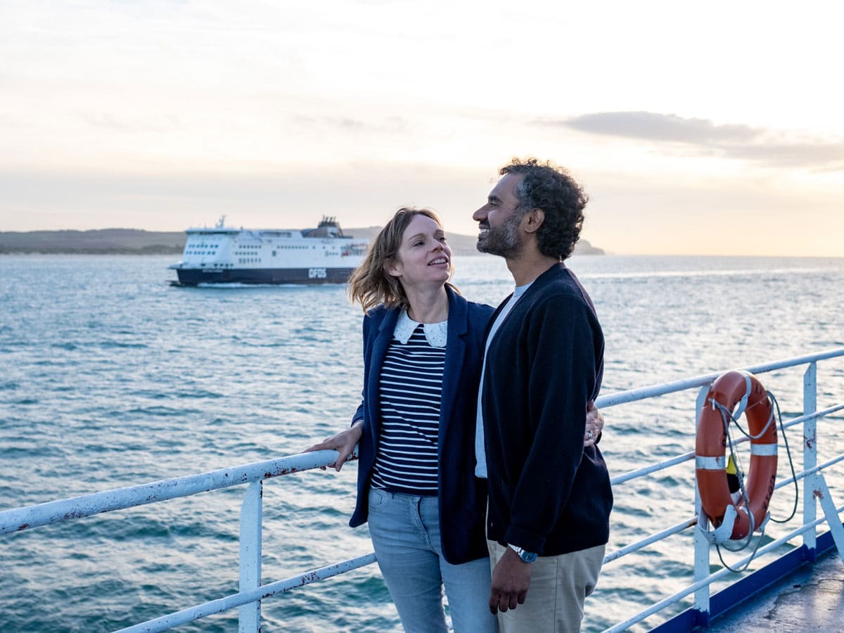 Ein Paar steht an der Reling einer Fähre und blickt über das Meer, während im Hintergrund eine weitere große Autofähre vorbeifährt.