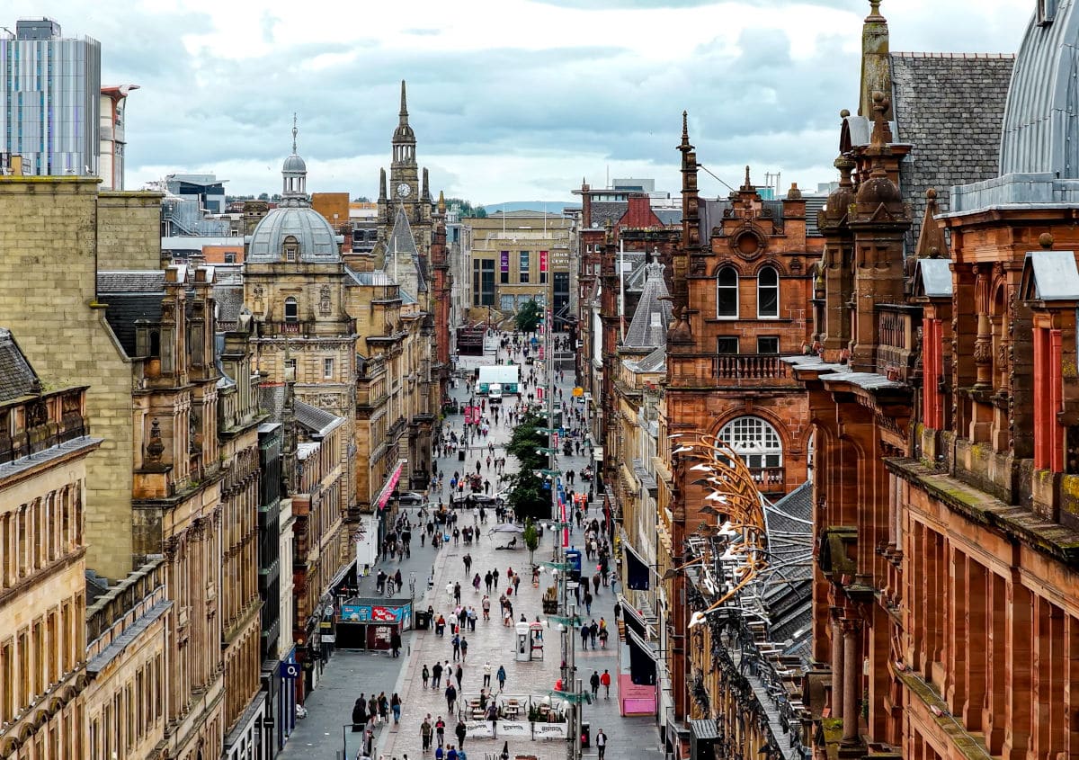 Die Aufnahme zeigt eine belebte Fußgängerzone im Zentrum von Glasgow, umgeben von historischen Sandsteingebäuden und imposanter viktorianischer Architektur. Viele Menschen schlendern die Einkaufsstraße entlang, während sich am Horizont markante Kirchtürme und moderne Bauten abzeichnen.