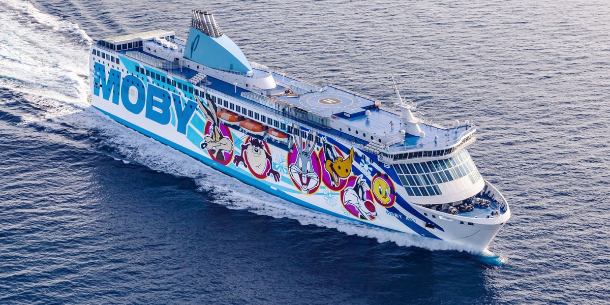 Auf dem Bild fährt eine große Autofähre mit dem Schriftzug „MOBY“ und bunten Comicfiguren wie Bugs Bunny, Tweety und Taz auf dem offenen Meer. Die Fähre hebt sich mit ihrer auffälligen Bemalung und dem modernen Design deutlich vom blauen Wasser ab.