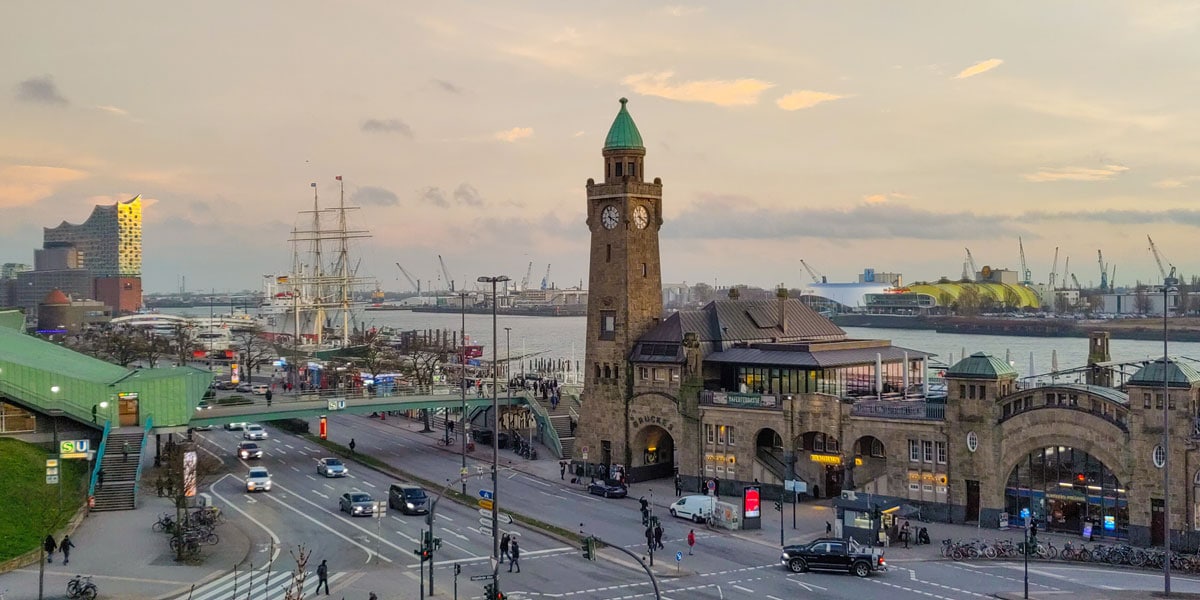 Ein belebter Blick auf die Hamburger Landungsbrücken mit der markanten Uhrturm-Gebäudeanlage im Vordergrund, mehrspuriger Straße und Fußgängern darunter, der Elbe mit Hafenkränen, Schiffen und der Elbphilharmonie im Hintergrund bei Abenddämmerung.