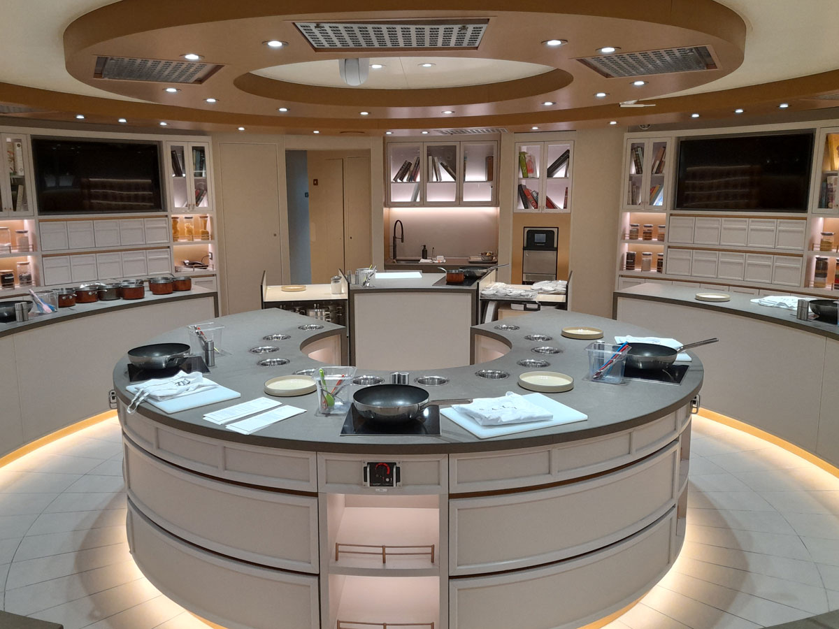 Modernes Kochstudio auf einem Kreuzfahrtschiff mit halbrunden Arbeitsflächen, mehreren Kochfeldern, Pfannen, Tellern und Kochutensilien; helle Beleuchtung und elegante Einrichtung schaffen eine einladende Atmosphäre für Kochkurse oder Showkochen auf See.