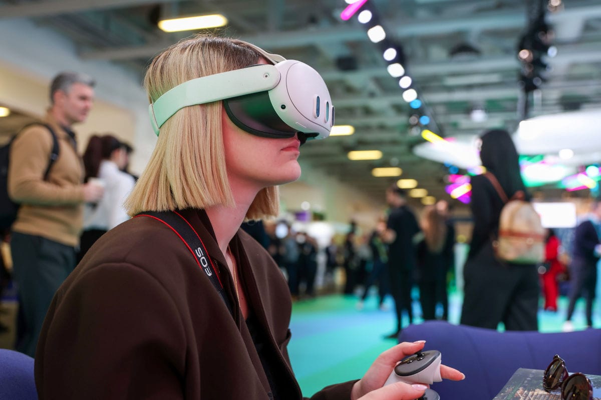 Eine Frau mit blonden Haaren trägt eine VR-Brille und hält einen Controller in der Hand, sitzt auf einem Sofa auf einer Messe mit unscharfen Besuchern und bunten Lichtern im Hintergrund.