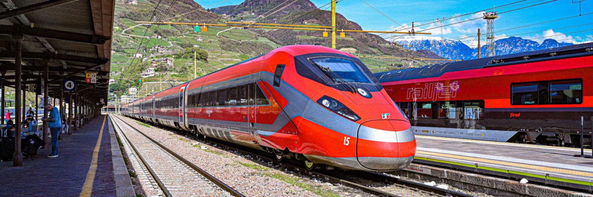 260220-trenitalia-roter-schnellzug-im-bahnhof Auf dem Bild ist ein moderner, roter Trenitalia-Schnellzug zu sehen, der im Bahnhof vor einer malerischen Bergkulisse steht. Am Bahnsteig warten Reisende neben Gleisen, während im Hintergrund ein weiterer Zug des Railjet zu erkennen ist. Das Foto zeigt italienischen Hochgeschwindigkeitszug, Zugverkehr und Bahnhofsatmosphäre in den Alpen.