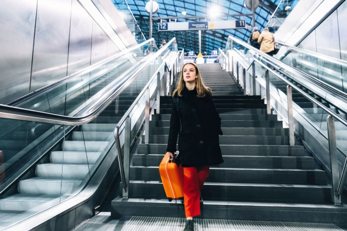 Eine Frau mit orangefarbenem Koffer geht eine Treppe im modernen Berliner Hauptbahnhof hinunter, flankiert von Rolltreppen und glänzenden Metallwänden; das Bild vermittelt die Atmosphäre von Reisen, Deutsche Bahn und Mobilität in Berlin. Im Hintergrund sind weitere Reisende sowie Bahnsteigschilder mit den Zahlen 11 und 12 sichtbar. Das Glasdach und die Architektur des Bahnhofs betonen den urbanen Charakter des Verkehrsknotenpunkts.