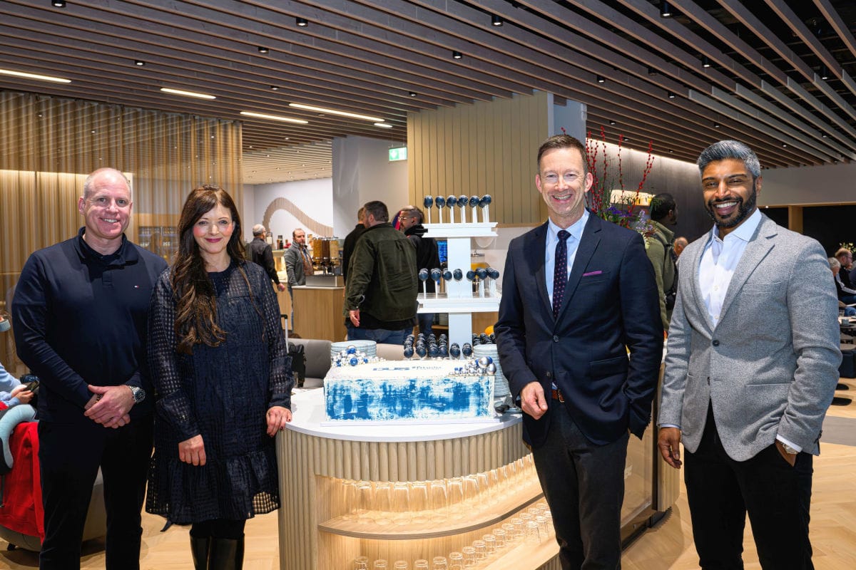 In der modernen Flughafen-Lounge Düsseldorf stehen vier festlich gekleidete Personen vor einem großen, blau-weißen Eröffnungskuchen und einer Etagere mit Cake Pops, während im Hintergrund weitere Gäste die stilvolle Einrichtung und das Ambiente genießen. Die Szene vermittelt die feierliche Eröffnung der neuen Lounge am Düsseldorfer Flughafen und unterstreicht das elegante Lounge-Design sowie die hochwertige Gastronomie.