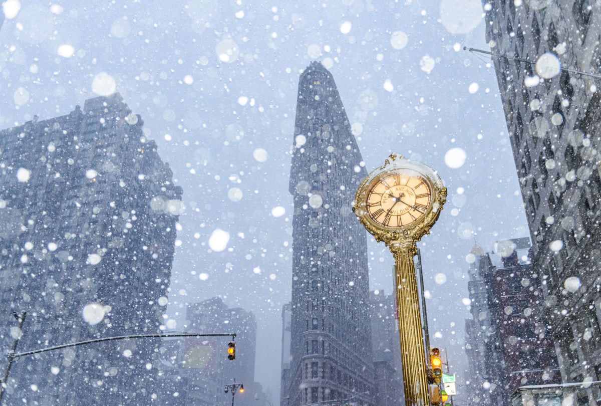 Das Bild zeigt einen heftigen Schneesturm in New York City mit großen Schneeflocken, die vor dem berühmten Flatiron Building und einer goldenen Straßenuhr auf der 5th Avenue herunterfallen. Die winterliche Szene vermittelt die Auswirkungen eines starken Schneefalls auf das Stadtbild von Manhattan.