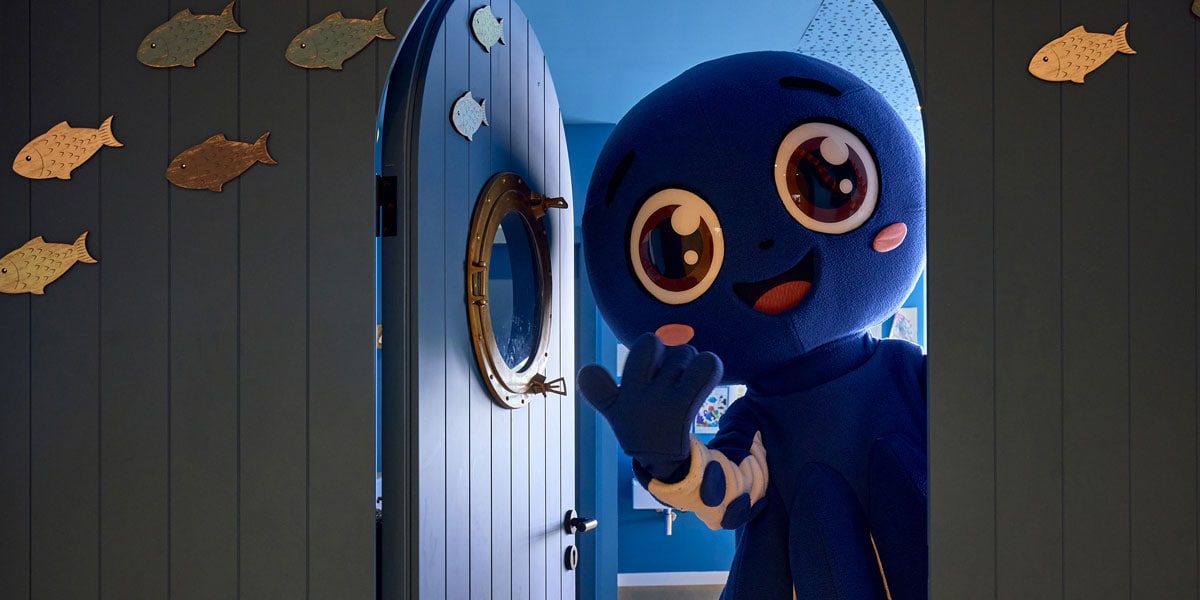 Blaues Maskottchen Pop winkt an einer Tür des Kids Sea Club mit Fisch-Dekoration und maritimer Gestaltung.