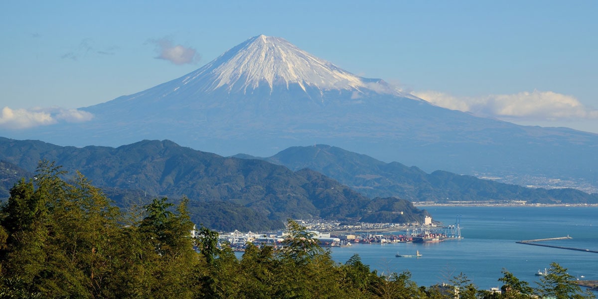 Auf dem Foto ist der schneebedeckte Berg Fuji in Japan zu sehen, der sich majestätisch über eine grüne Hügellandschaft erhebt. Im Vordergrund liegt ein Hafen am blauen Meer, während klare Sicht den berühmten Vulkan deutlich erkennen lässt.