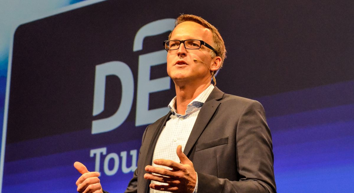 Ein Mann in Anzug und Hemd steht auf einer Bühne vor einer groß projizierten Präsentationsfolie und spricht gestikulierend ins Publikum.