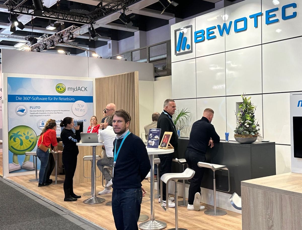 Messestand von Bewotec auf einer Fachmesse mit großem Firmenlogo an der Wand. Mehrere Personen stehen an Hochtischen und am Counter im Gespräch. Links ist ein „myJACK“-Banner mit der Aufschrift „Die 360°-Software für Ihr Reisebüro“ zu sehen. Moderne Standgestaltung mit hellen Holz- und Weißtönen sowie Blumendekoration auf dem Tresen.