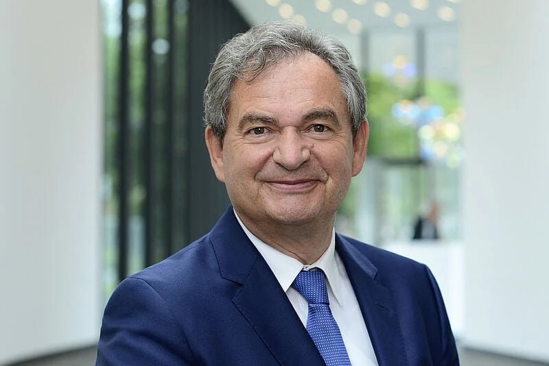 Ein Mann im blauen Anzug mit weißem Hemd und blauer Krawatte steht in einem modernen, lichtdurchfluteten Bürogebäude. Im Hintergrund sind große Fenster, grüne Pflanzen und unscharfe Lichter zu sehen.