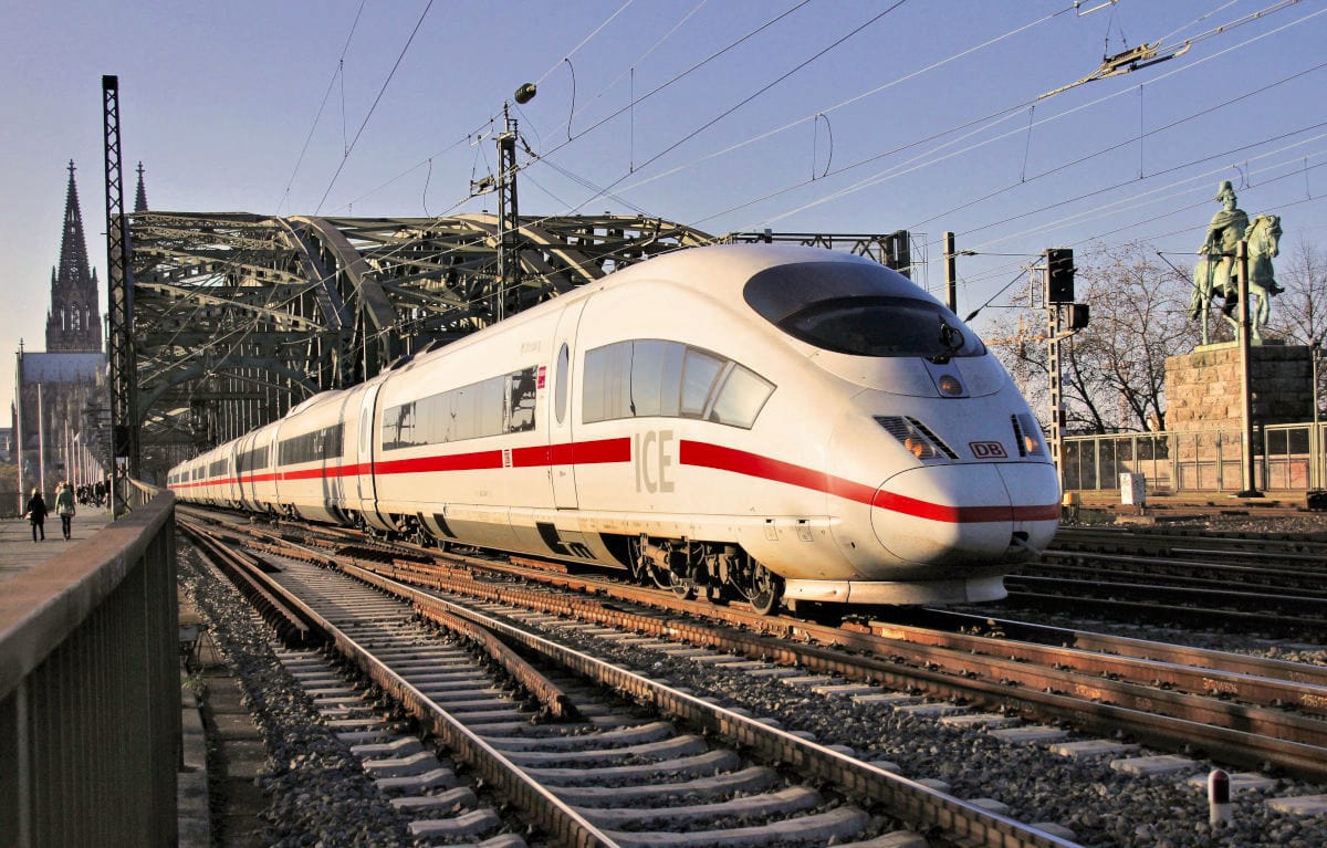 Ein moderner ICE-Zug fährt bei Sonnenschein über die Hohenzollernbrücke in Köln, im Hintergrund ist der Kölner Dom zu sehen. Das Foto zeigt den schnellen Intercity-Express auf den Gleisen, umgeben von markanten Wahrzeichen der Stadt.