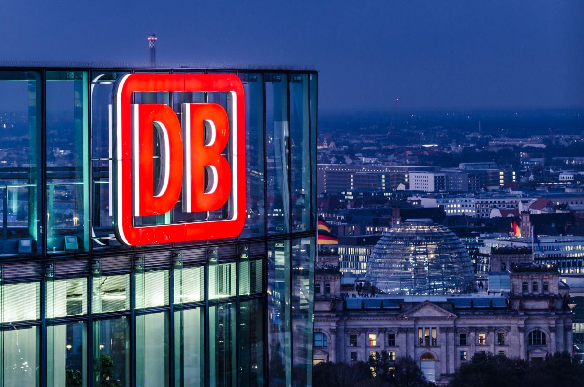 Das Bild zeigt das beleuchtete Logo der Deutschen Bahn auf einem modernen Glasgebäude in Berlin bei Nacht, im Hintergrund ist die Kuppel des Reichstagsgebäudes zu sehen. Das Foto verbindet die Themen Deutsche Bahn, Konzernzentrale und Berlin bei Nacht.