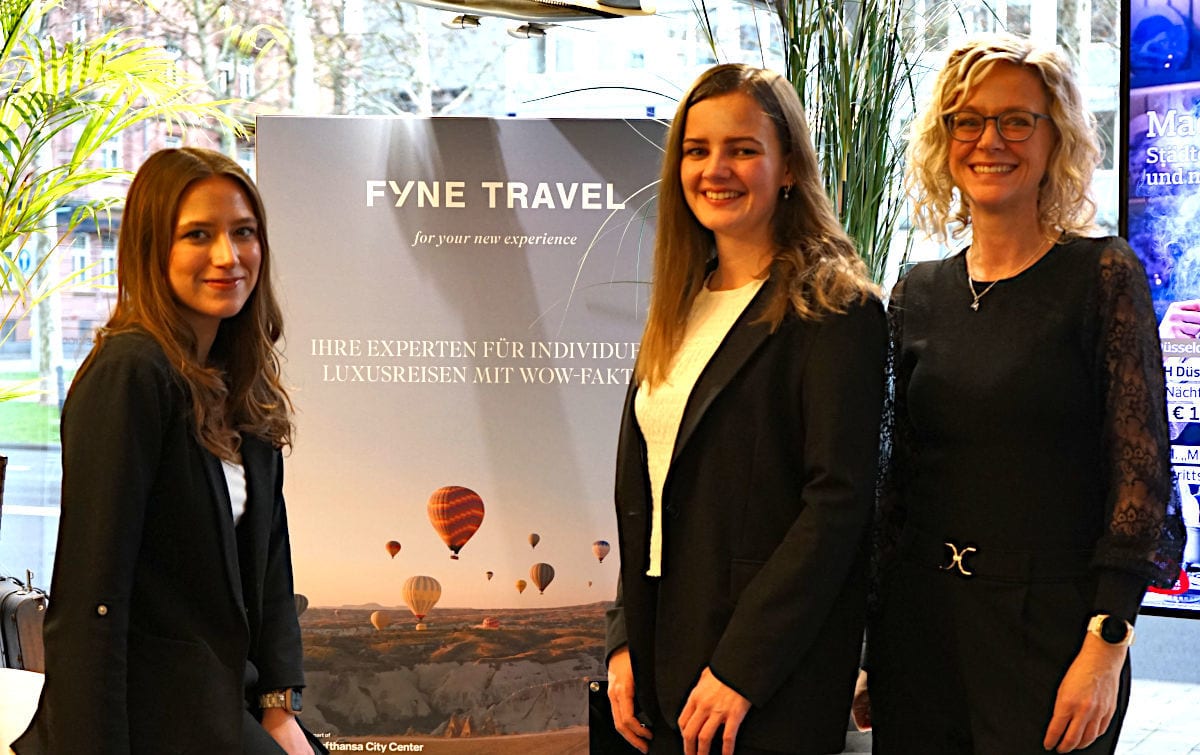 Drei Frauen in dunkler Business-Kleidung stehen in einem Reisebüro vor einem Fyne-Travel-Poster mit Heißluftballons und lächeln in die Kamera; im Hintergrund sind große Fenster und Pflanzen zu sehen.
