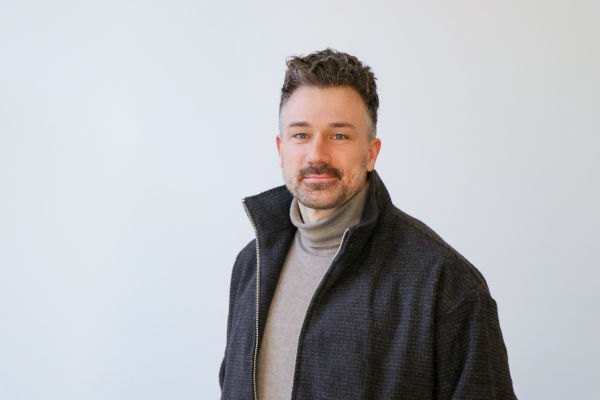 Porträt eines Mannes mit kurzen, leicht gewellten dunklen Haaren und Bartstoppeln, der vor hellem, neutralem Hintergrund steht. Er trägt einen beigen Rollkragenpullover und eine dunkle Jacke mit Reißverschluss und blickt ruhig in die Kamera.