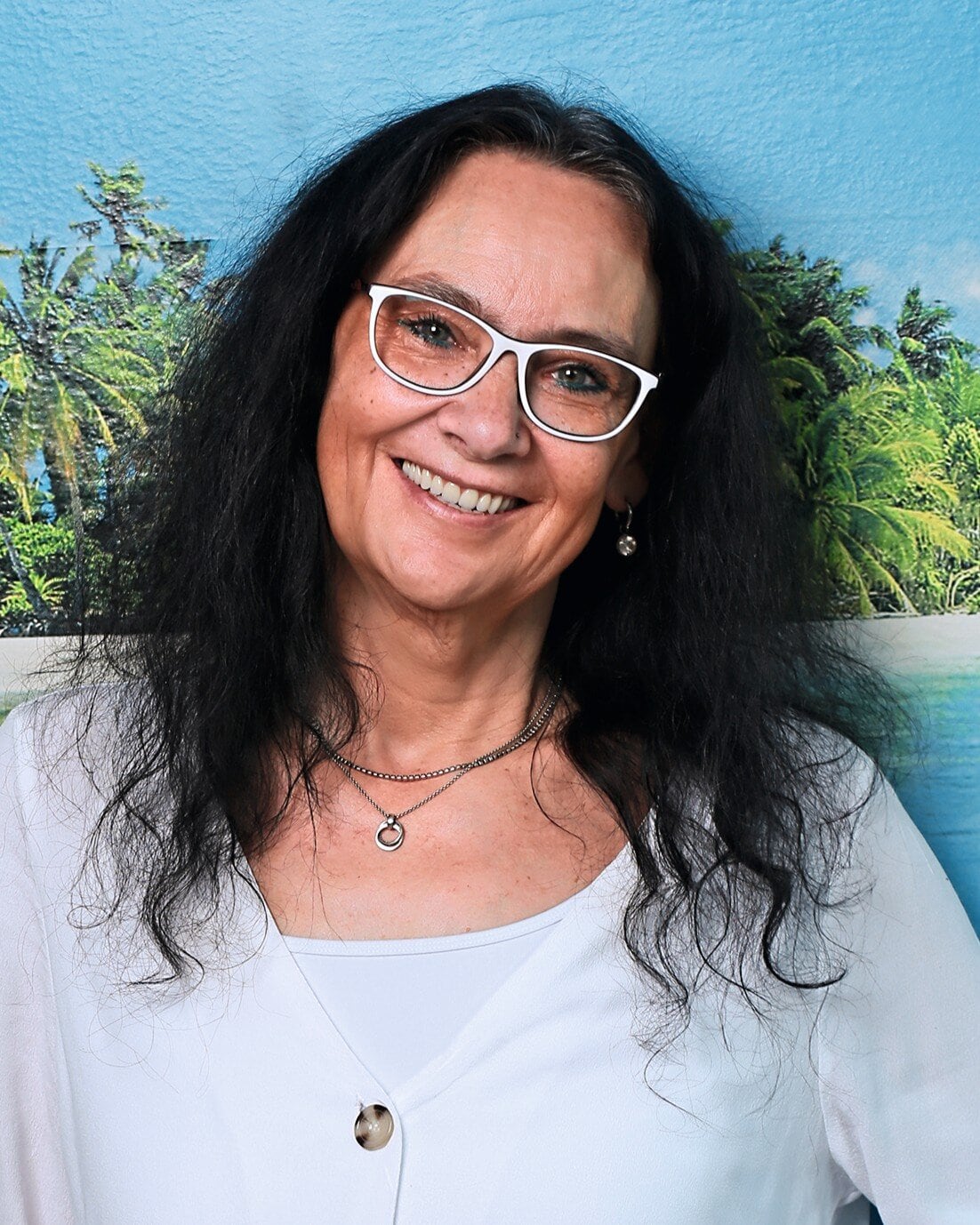 Eine Frau mit schwarzen langen Haaren und weiß-umrandeter Brille steht vor einem Urlaubsfoto und lacht