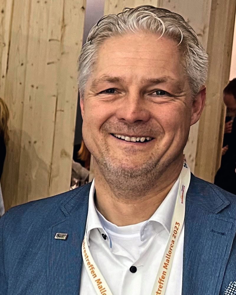 Ein Mann vor einer Holzwand mit grauen Haaren und blauem Blazer lacht in die Kamera