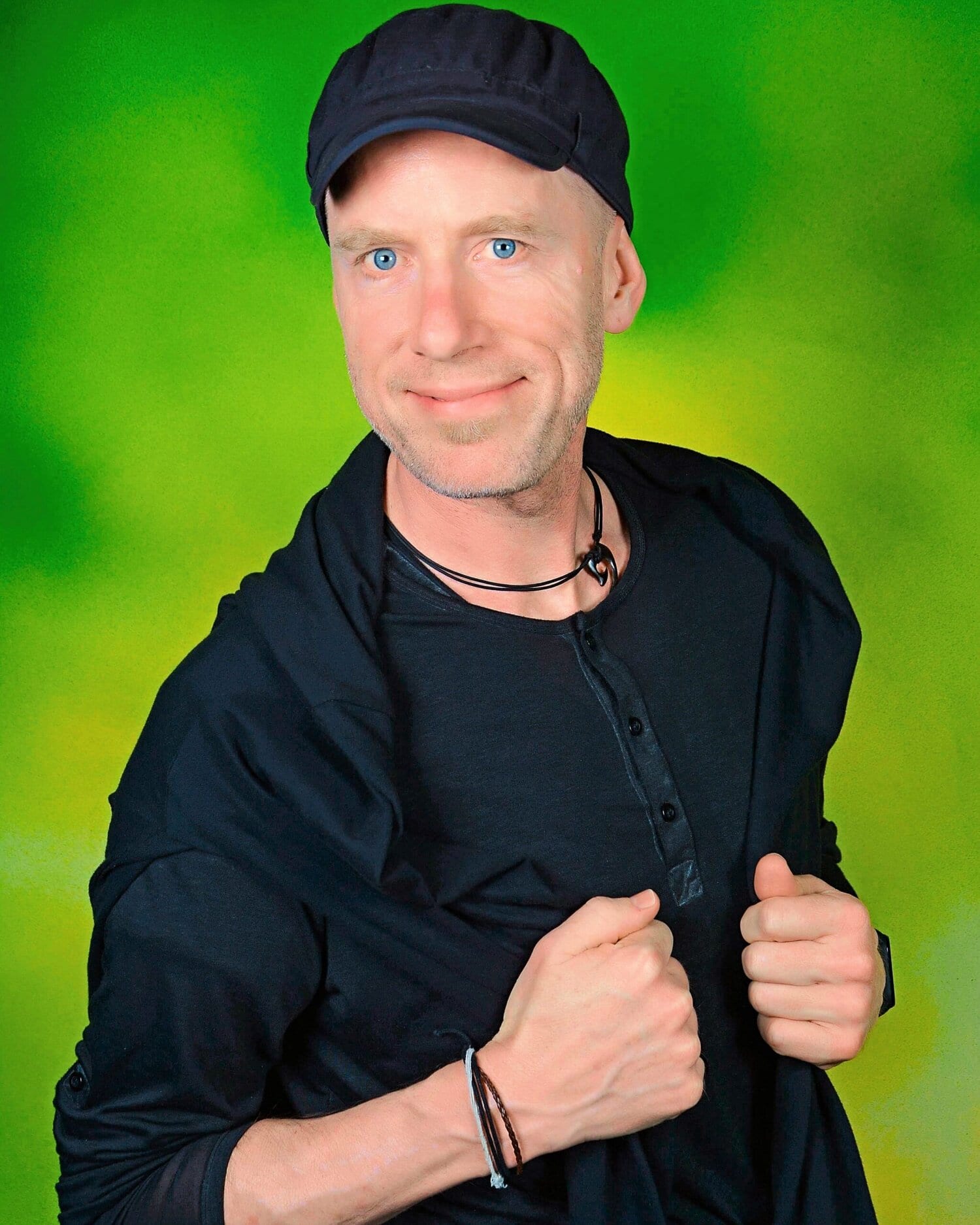 Ein schwarzgekleideter Mann mit schwarzer Kappe vor grünem Hintergrund lächelt in die Kamera