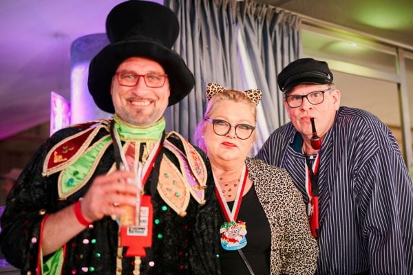 Drei Menschen in fantasievollen Karnevalskostümen posieren gemeinsam bei einer Karnevalsparty, einer trägt einen Zylinderhut und bunte Jacke, eine andere ein Leopardenkostüm. Die Szene ist lebendig und vermittelt ausgelassene Karnevalsstimmung. Im Hintergrund sieht man weitere Gäste und festliche Dekoration.