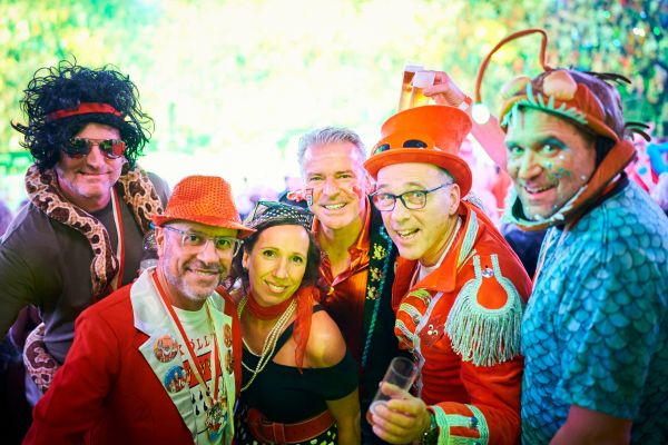 Sechs Karnevalisten in bunten und kreativen Kostümen feiern gemeinsam, darunter auffällige Hüte, Perücken und Tierkostüme. Die Gruppe steht eng beisammen und lacht, während die festliche Beleuchtung für Partyatmosphäre sorgt. Karneval, Kostüme und Gruppenspaß machen das Bild aus.