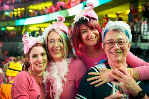 Vier Menschen feiern verkleidet beim Karneval, drei tragen pinke Flamingohüte und Federboas, einer ist als Matrose verkleidet. Die Gruppe steht in einer festlich beleuchteten Halle voller Menschen. Karneval, Kostüme und ausgelassene Partylaune prägen das Bild.