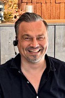 Ein Mann mit Headset und schwarzem Hemd lacht in die Kamera