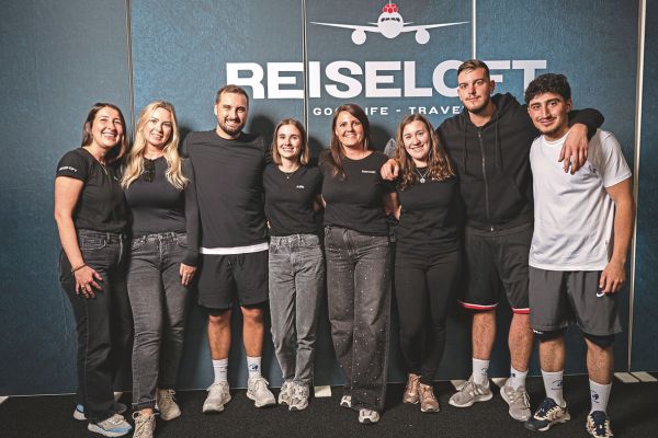 Eine Gruppe von jungen Menschen steht vor einer grauen Wand mit Reiseloft. Sie legen sich die Hände um die Schultern und lachen