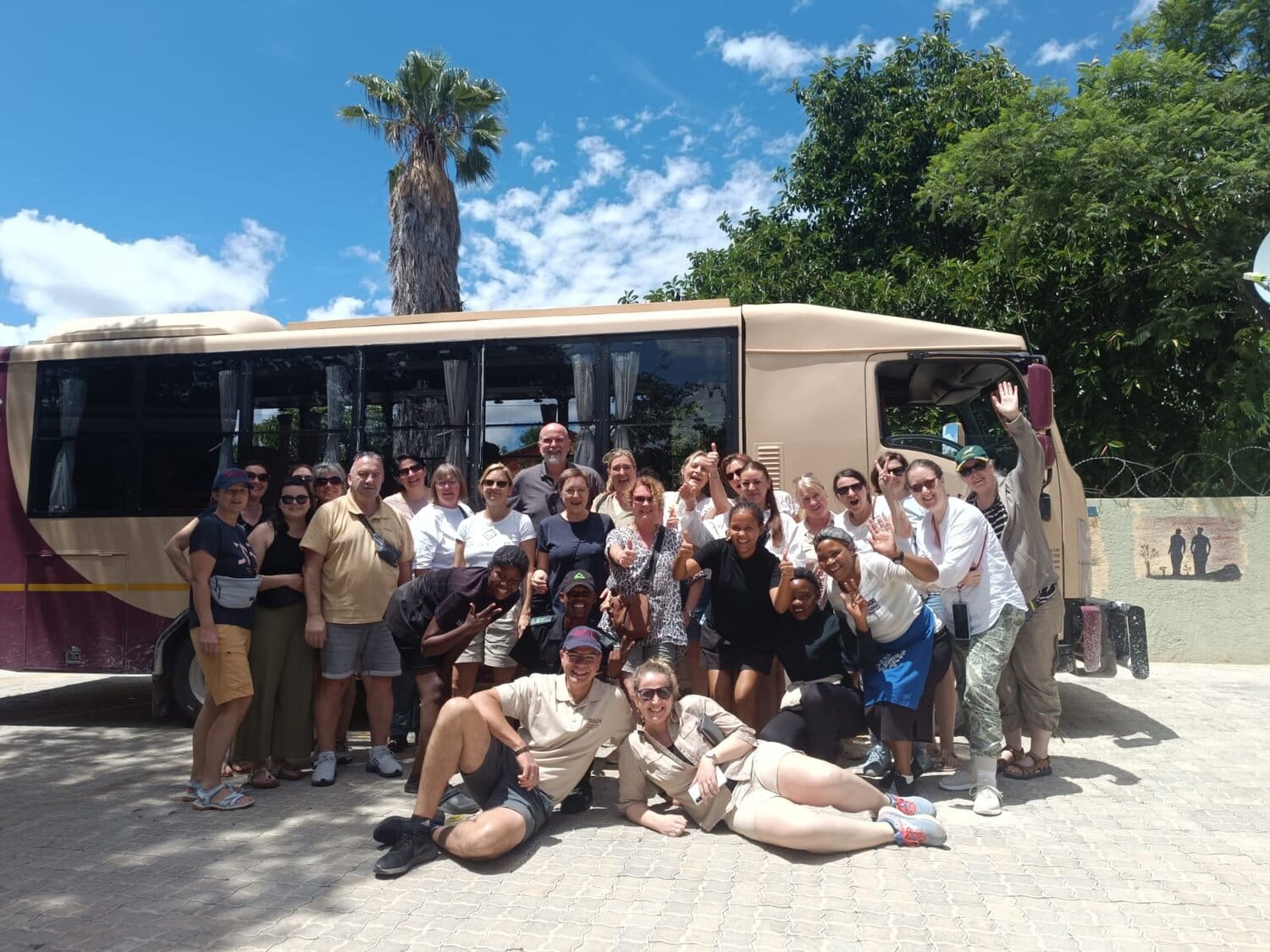Eine Gruppe von etwa 20 fröhlichen Menschen posiert vor einem beigen Reisebus unter blauem Himmel mit Palmen im Hintergrund; zwei Personen liegen entspannt vorne auf dem Boden, während die anderen dicht dahinter stehen und in die Kamera lächeln oder winken.