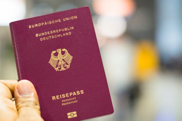 Eine Hand hält einen bordeauxroten deutschen Reisepass mit goldener Prägung („Europäische Union“, „Bundesrepublik Deutschland“, Bundesadler) vor unscharfem Hintergrund, der an einen Flughafen oder Check-in-Bereich erinnert.