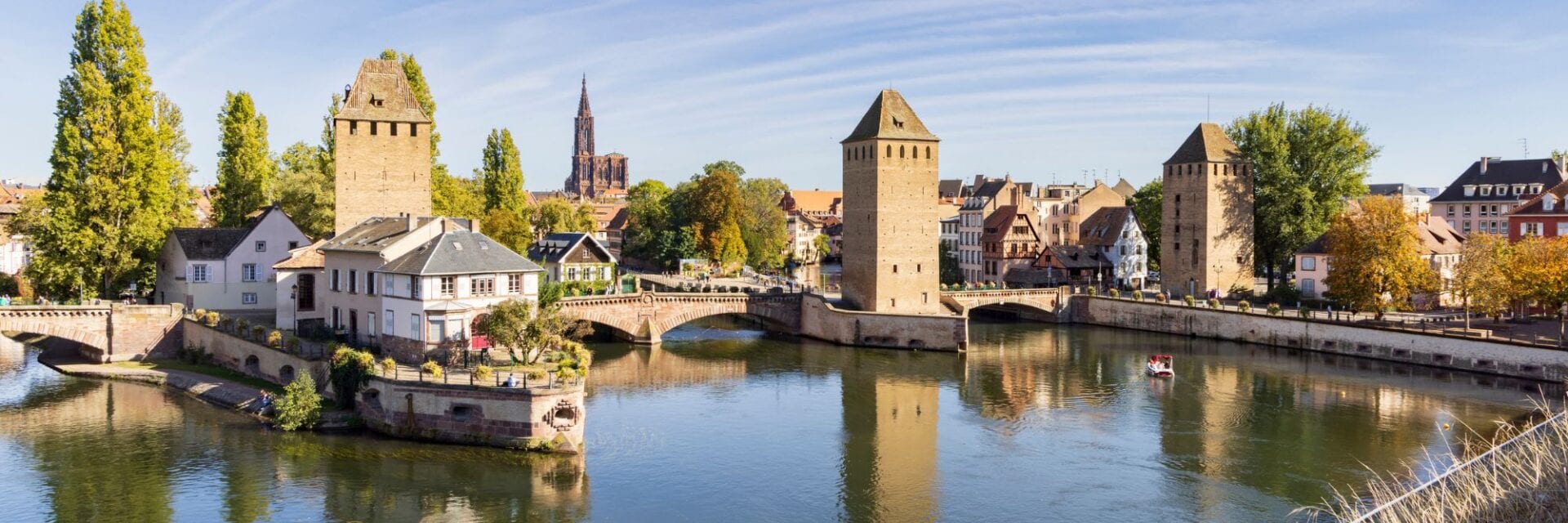 Panoramablick auf Straßburg mit historischen Brückentürmen und Fachwerkhäusern am Fluss; im Hintergrund ragt das Münster über die Altstadt, Bäume spiegeln sich im ruhigen Wasser.