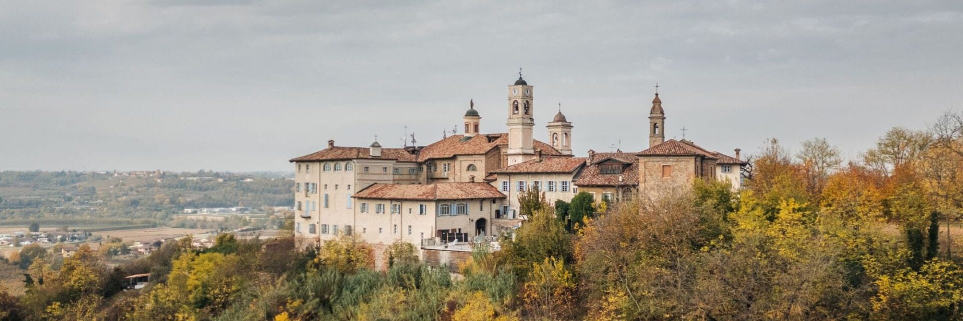 Das Foto zeigt das historische Borgo Monchiero Resort in Italien, eingebettet in herbstlich gefärbte Weinberge der Langhe-Region. Die malerische Schlossanlage mit ihren charakteristischen Türmen und roten Ziegeldächern erhebt sich über die sanfte Hügellandschaft.