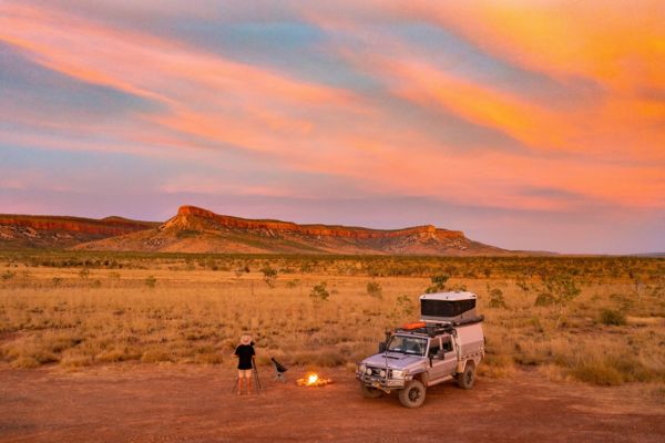 Allrad-Camper mit Dachzelt in der australischen Outback-Landschaft der Kimberley-Region bei Sonnenuntergang; eine Person fotografiert neben einem kleinen Lagerfeuer vor roten Felsformationen unter farbenprächtigem Himmel.