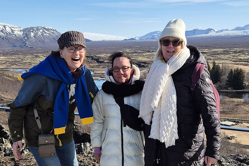 Drei warm eingepackte Personen posieren im Vordergrund einer weiten, offenen Landschaft mit Fluss, braunen Wiesen und schneebedeckten Bergen unter blauem Himmel; sie tragen Winterjacken, Schals und Mützen, die Gesichter sind unkenntlich gemacht.
