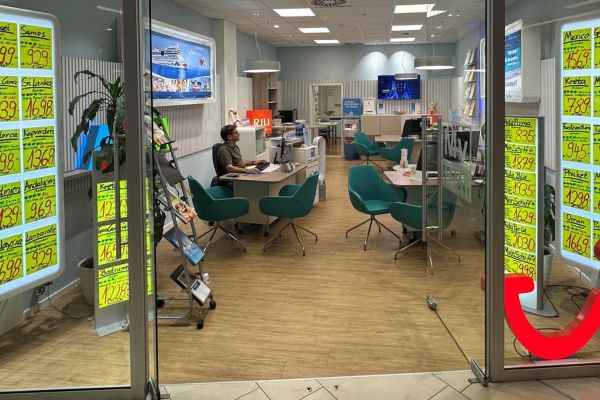 Reisebüro in einem Einkaufszentrum mit großer Glasfront; an den Fenstern hängen zahlreiche leuchtend gelbe Angebotsplakate mit Reisezielen und Preisen. Im Inneren sitzt ein Mitarbeiter an einem Schreibtisch, umgeben von modernen türkisfarbenen Stühlen und Beratungstischen. Auf der rechten Glasscheibe ist ein rotes, lächelndes Logo zu sehen.