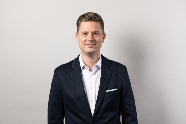 Porträt eines Mannes mit kurzen, hellbraunen Haaren in dunklem Anzug und weißem Hemd, vor neutral grauem Hintergrund, freundlich in die Kamera blickend.