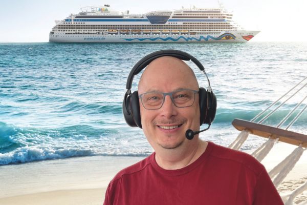 Ein lächelnder Mann mit Brille und Headset steht vor einem Strand mit türkisblauem Meer; im Hintergrund fährt ein großes Kreuzfahrtschiff vorbei.