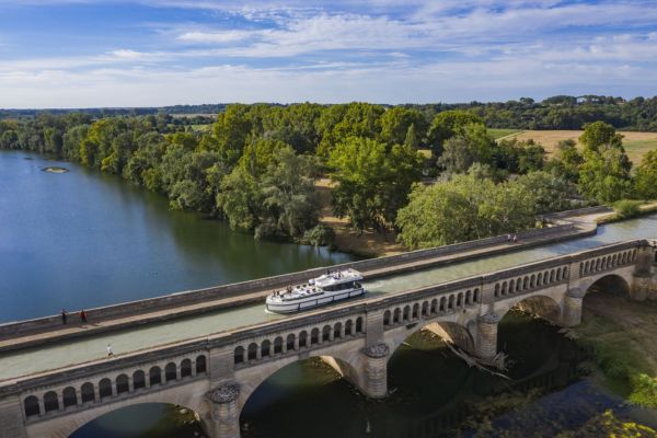 Luftaufnahme eines weißen Hausboots, das über einen historischen steinernen Bogenbrücken-Kanal (Pont-Canal du Canal du Midi) fährt; darunter fließt ein breiter Fluss mit üppigen grünen Uferbäumen, Feldern und einer kleinen Insel unter blauem Himmel mit Wolken.