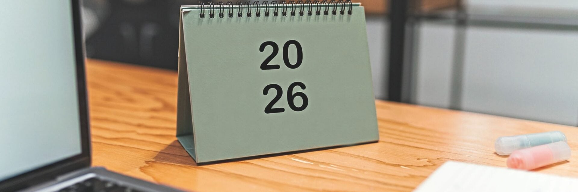 Ein aufgestellter Tischkalender mit Spiralbindung zeigt die Jahreszahl 2026 und steht auf einem Holztisch; daneben liegen ein Laptop und zwei Textmarker.