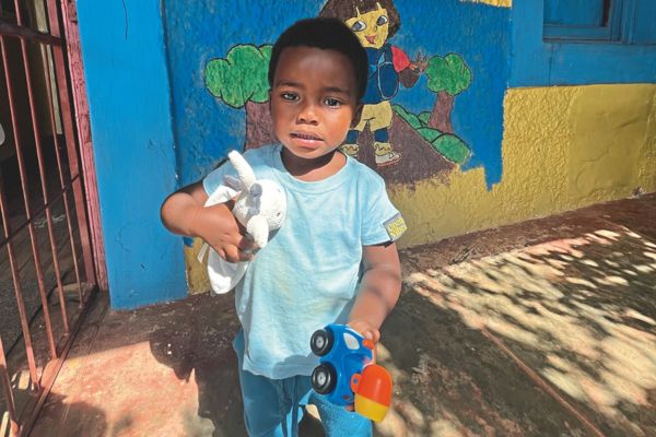 Ein Kleinkind steht barfuß vor einem farbenfroh bemalten Kindergarten in Südafrika und hält ein Stofftier und ein Spielzeugauto in den Händen.