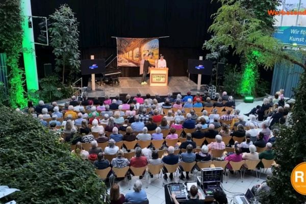 Blick von oben auf einen Saal, in dem viele Menschen sitzen und auf eine Bühne schauen