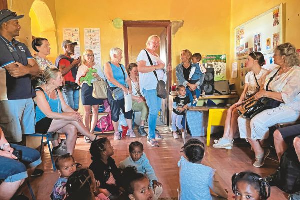 Besucherinnen und Besucher stehen und sitzen gemeinsam mit Kleinkindern in einem Kindergartenraum in Südafrika und hören einer Frau zu, die in der Mitte spricht.