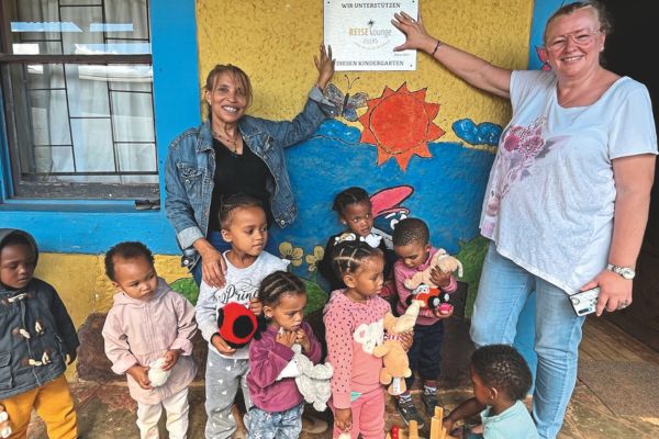 Kinder eines südafrikanischen Kindergartens stehen vor einem bunt bemalten Gebäude, flankiert von zwei erwachsenen Frauen, die auf eine Unterstützerplakette an der Wand zeigen.