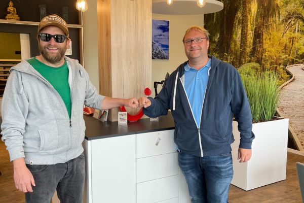 Zwei Männer stehen lächelnd am Counter eines Reisebüros und begrüßen sich mit einem Fistbump; moderne Einrichtung und ein großflächiges Naturmotiv prägen den Raum.