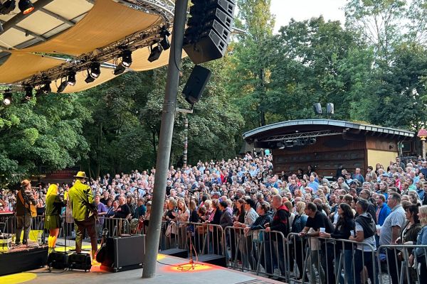 Publikum vor einer Open-Air-Bühne bei einem Konzert im Grünen; Musiker stehen unter dem Bühnendach, zahlreiche Besucher verfolgen die Live-Performance dicht gedrängt vor der Bühne.