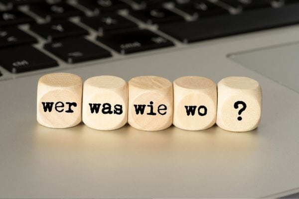 Fünf Holzklötze mit der Aufschrift „wer was wie wo ?“ liegen nebeneinander auf einer Laptop-Tastatur und symbolisieren zentrale W-Fragen.