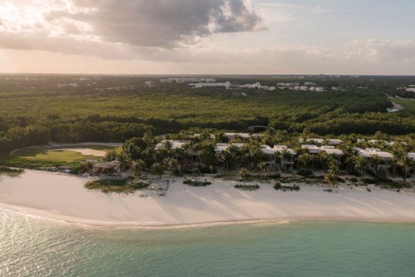 Das Foto zeigt das luxuriöse Hyatt Alila Mayakoba Resort an der Riviera Maya in Mexiko, eingebettet in üppige grüne Vegetation direkt an einem langen weißen Sandstrand mit türkisfarbenem Meer. Im Hintergrund erstreckt sich dichter Dschungel, während das Resort inmitten von Palmen und tropischen Pflanzen liegt.