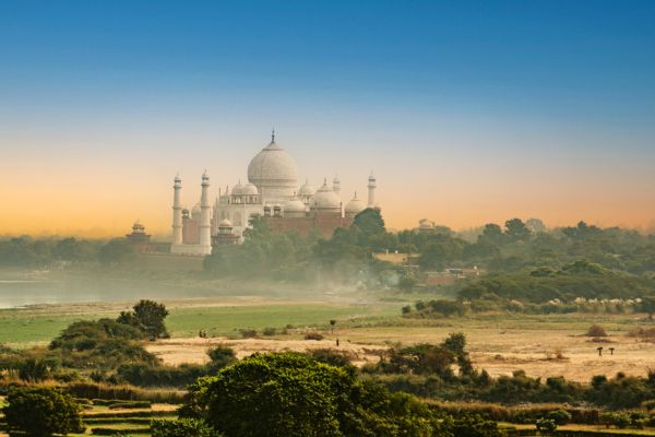 Blick auf das Taj Mahal im Morgenlicht, umgeben von Nebel und grüner Landschaft unter einem klaren Himmel.