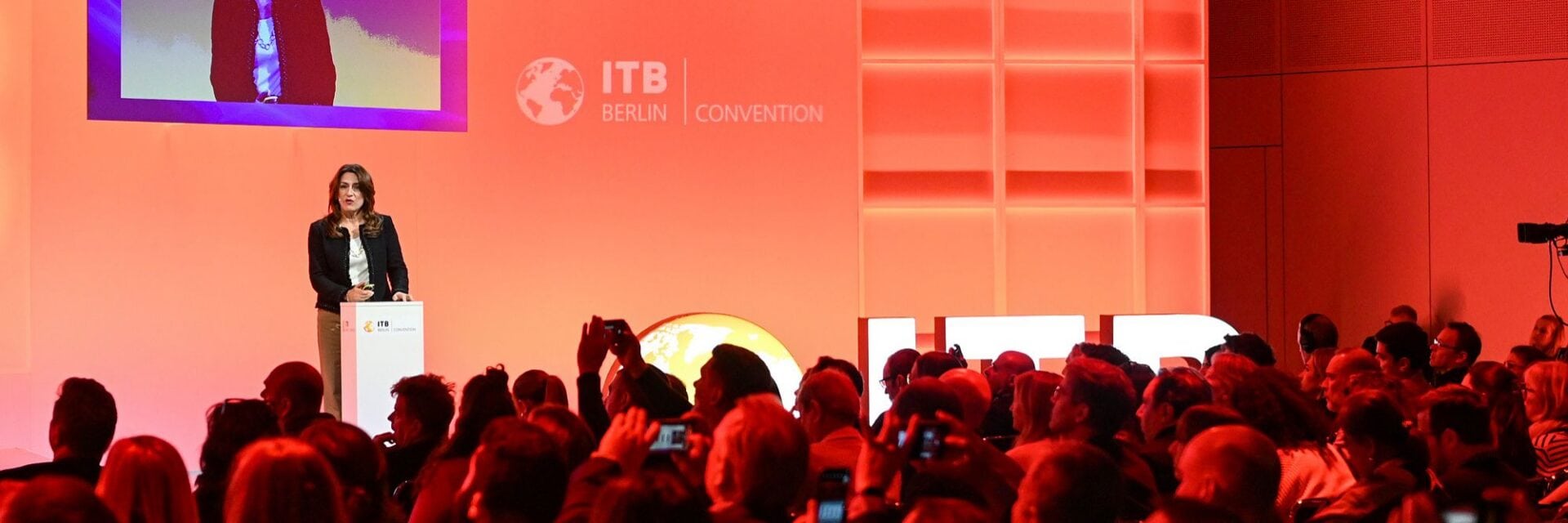 Ein Konferenzsaal mit vielen sitzenden Zuhörerinnen und Zuhörern, vor ihnen eine rot ausgeleuchtete Bühne mit einer stehenden Vortragenden; hinter ihr eine große Leinwand mit der Aufschrift „ITB Berlin Convention“ und Präsentationsfolien.