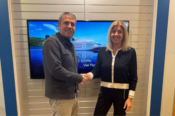 Ein Mann und eine Frau stehen vor einem Bildschirm mit einem Kreuzfahrtschiff-Motiv und schütteln sich die Hand. Beide lächeln, die Szene wirkt wie eine offizielle Übergabe oder geschäftliche Vereinbarung in einem modernen Büro.